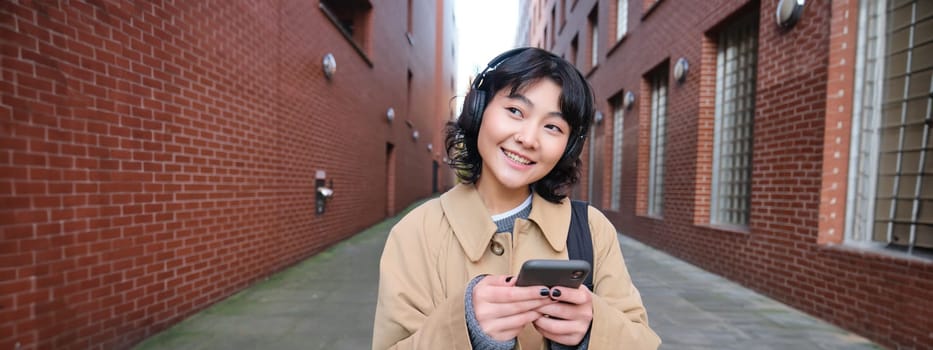 Portrait of young korean woman walking around city with backpack and headphones, listens music, looks at smartphone, uses phone application on streets.