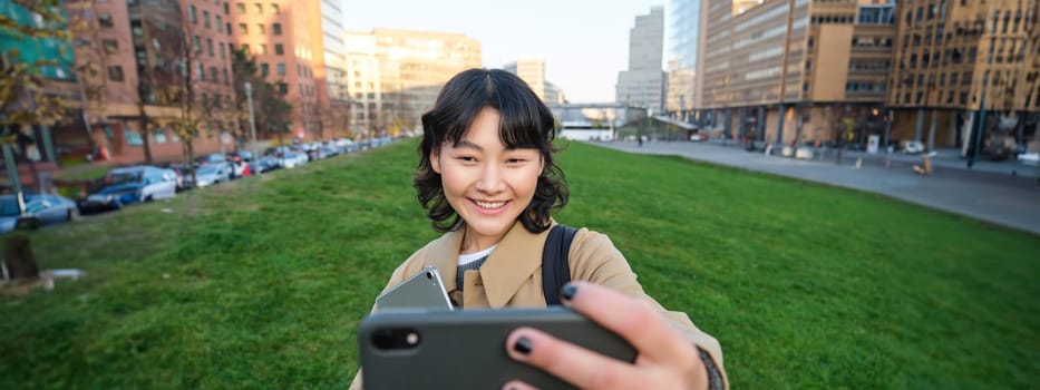 Happy young woman, student takes selfie on street, holds her tablet and homework notes, uses smartphone. Copy space