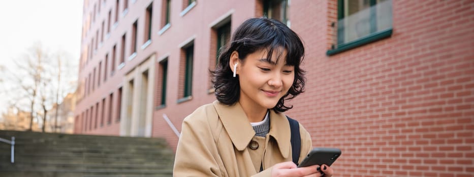 Happy korean girl walks on street, listens music in wireless earphones and holds smartphone, picks song in playlist while standing outdoor near building, reading message.