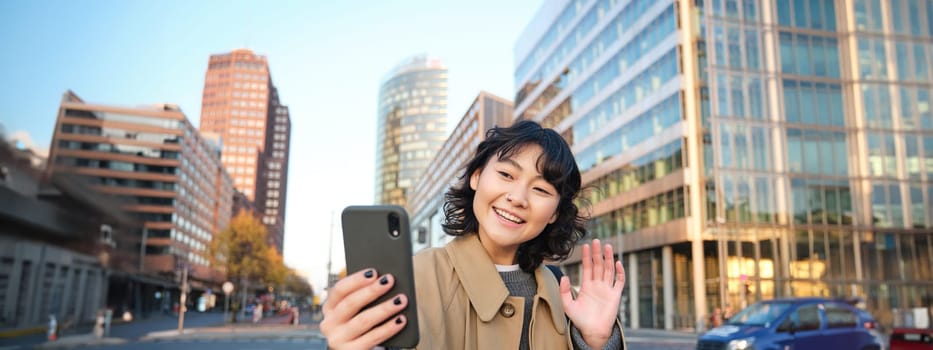 Happy asian girl tourist, takes selfie in city centre, makes video call and waves at smartphone camera, greets someone over phone.