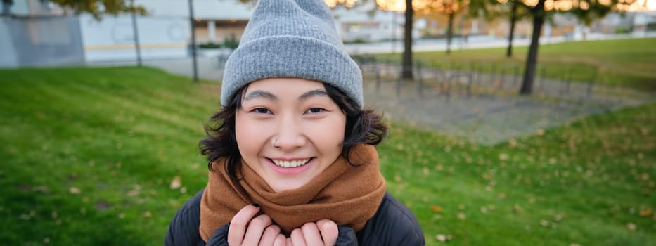 Portrait of cute asian girl in hat and scarf, walks around town in chilly spring weather, smiles and looks happy, sits in park near green grass.