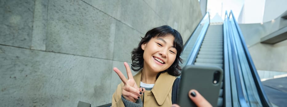 Positive asian girl in trench, shows peace sign, takes selfie while goes down escalator, smiles and looks happy.