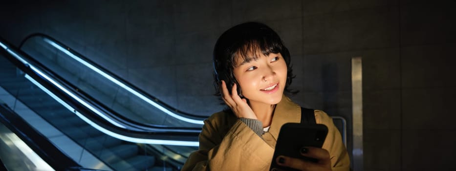 Cute young student, girl in headphones, commuting, standing near escalator, holding smartphone, listening music or podcast while travelling in city.