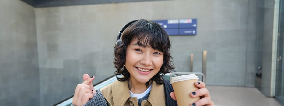 Portrait of stylish asian woman in headphones, drinks coffee to go and smiles, enjoys cappuccino while commutes, stands on street.