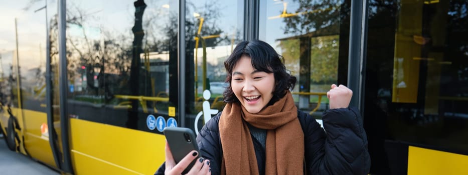 Portrait of cheerful asian girl talks on mobile phone, video chats, looks amazed at smartphone camera, stands on bus stop.