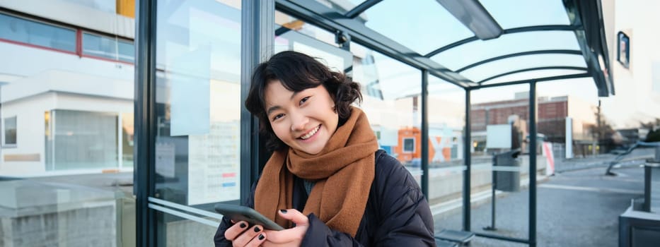 Cute smiling asian girl standing on bus stop, holding smartphone, wearing winter jacket and scarf. Woman commuting to work or university via public transport, stands on road.