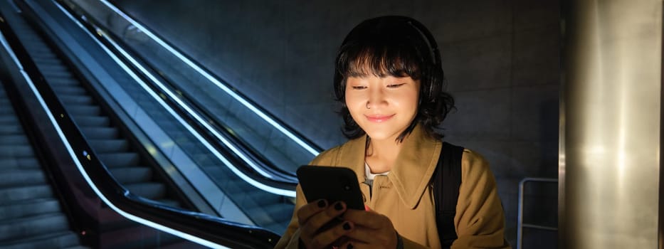 Smiling korean woman in headphones, reading message on smartphone, using mobile phone application, watching video or listens to music in public place.