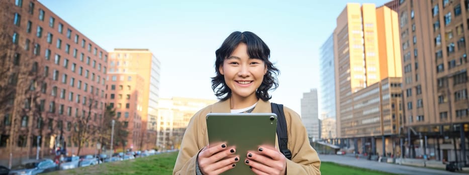 Happy young brunette girl, asian woman walks around city with tablet, goes to university with her digital gadget and backpack.