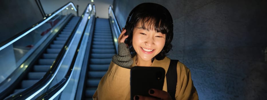 Beautiful young asian woman, going down an escalator, using smartphone, listening music in headphones while commuting to work on public transport.