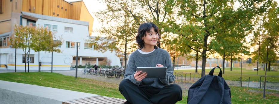 Young girl, korean artist or art student sits in park with digital tablet, draws with graphic pen, scatches a design or project, looks around.