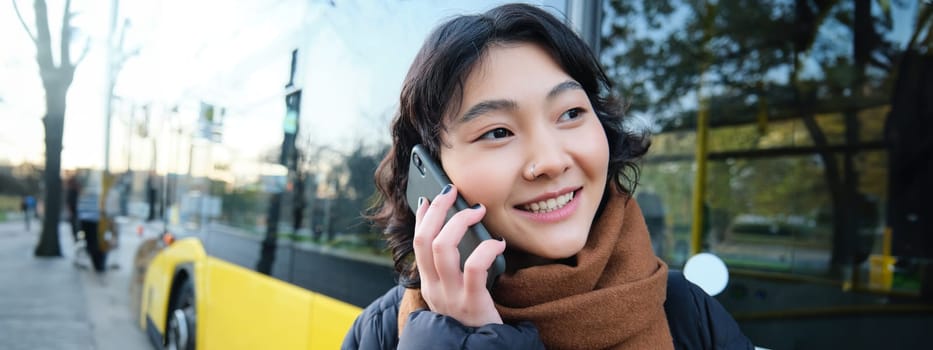 Cellular technology and people concept. Stylish asian girl talks on mobile phone, makes a telephone call, stands near bus stop and has conversation. Copy space