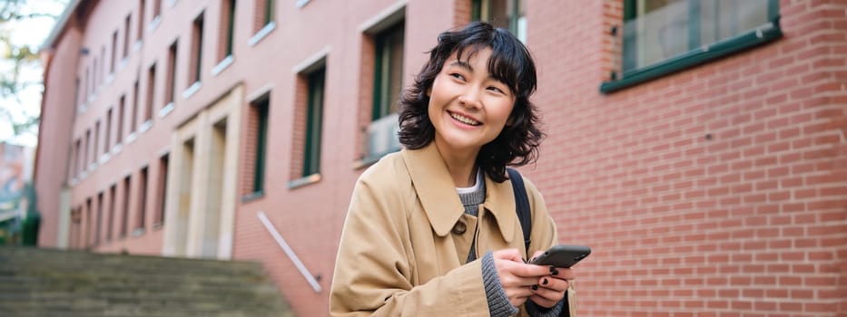 Happy korean girl walks on street, listens music in wireless earphones and holds smartphone, picks song in playlist while standing outdoor near building, reading message.