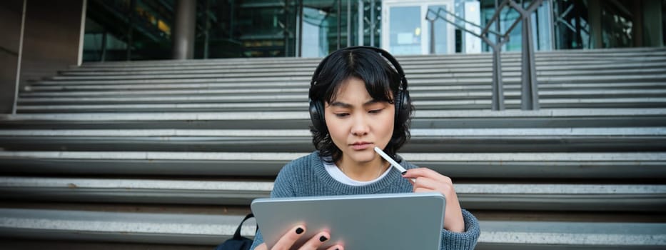 Portrait of asian girl with troubled face, draws on digital tablet with pen, looks complicated at her work, feels stuck no inspiration, sits on stairs and thinks.