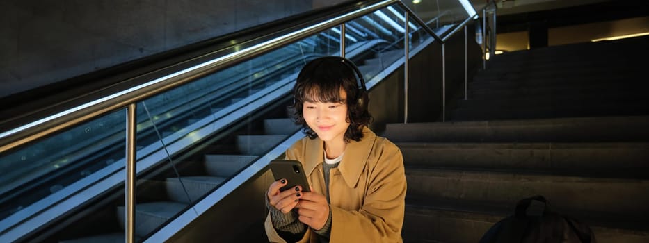 Technology. Portrait of asian girl sits on stairs near escalator, listens music in headphones and uses social media, reads, watches video in public place.