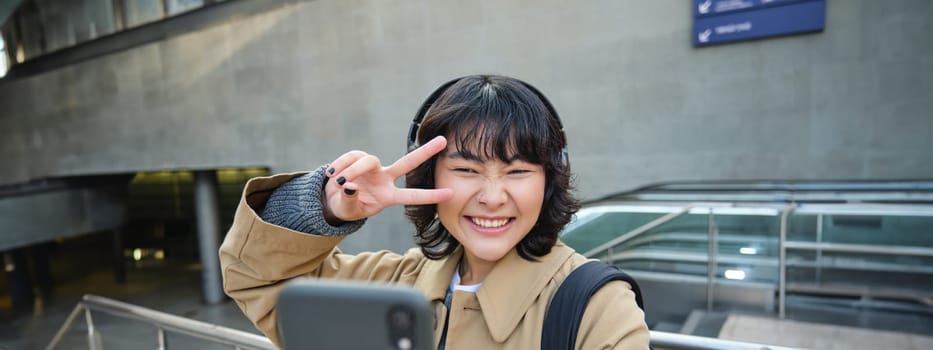 Beautiful korean girl in headphones, takes selfie on her smartphone, stands on street with mobile phone, records video, listens music and travels around city.