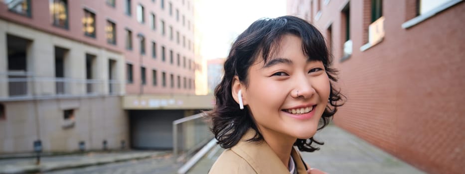 Walking around city. Smiling stylish korean girl in earphones, listens to music while walks on street, uses smartphone app, play songs.