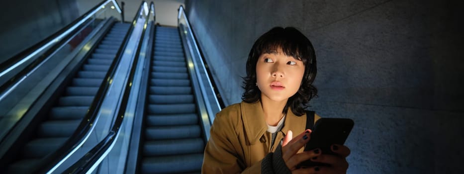 Urban lifestyle and people. Portrait of Korean girl in headphones, holds smartphone, looks concerned and tensed while going down escalator in dark.