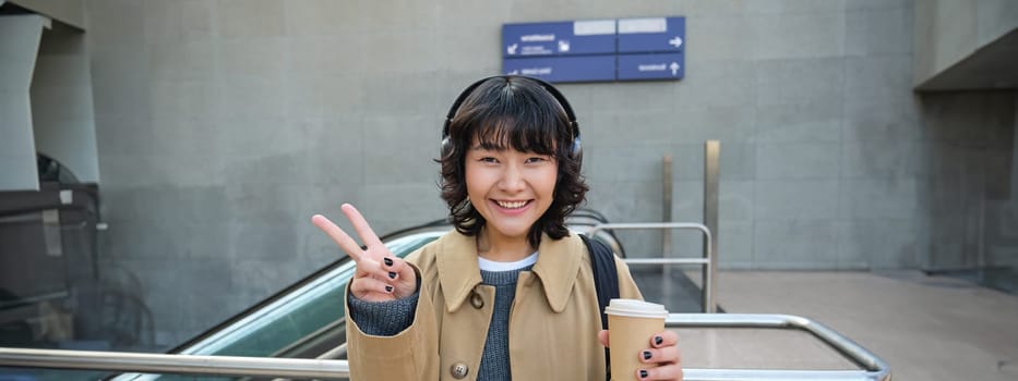Positive korean girl in headphones, drinks coffee to go, shows peace, v-sign, stands on street and smiles happily.