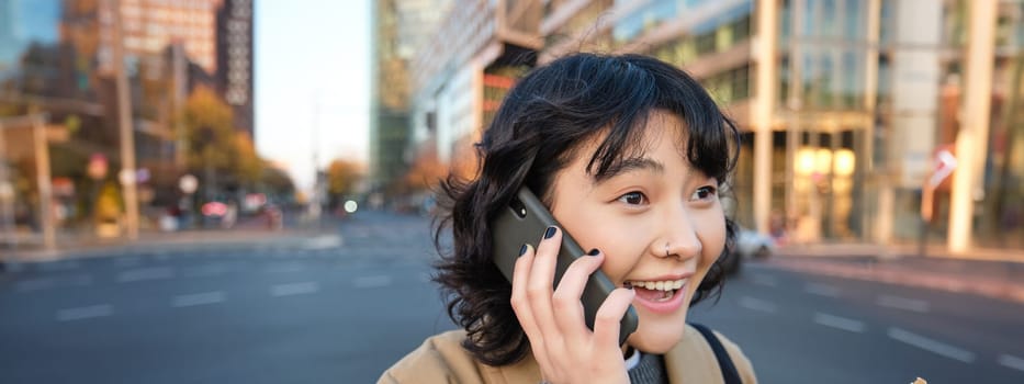 Portrait of surprised korean girl, talks on mobile phone, hears amazing news over telephone conversation, stands on streets of city with excited face expression.