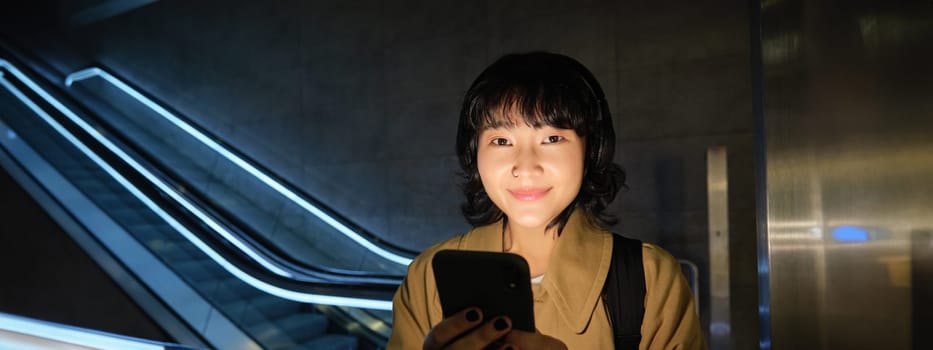 Happy asian woman in subway, standing near escalator, holding smartphone and listening music in headphones, smiling pleased.