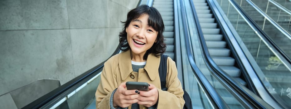Stylish korean woman, student on street, goes down escalator, holds smartphone and smiles cheerful at camera.