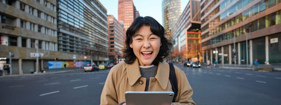 Enthusiastic asian girl with digital tablet, smiling and looking amazed, happy with something good, holding digital tablet, standing on street.