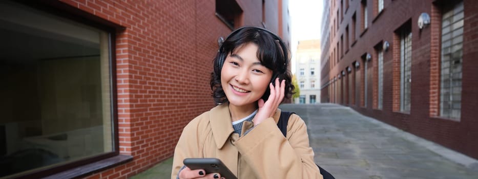 Portrait of young korean woman walking around city with backpack and headphones, listens music, looks at smartphone, uses phone application on streets.