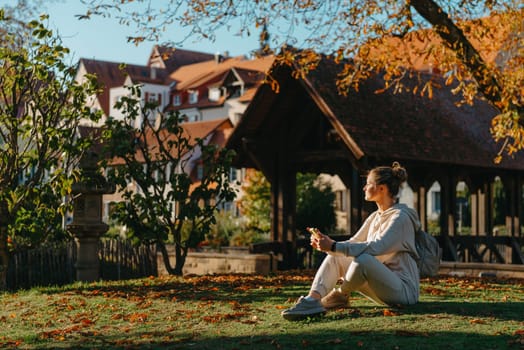 Young fashionable teenage girl with smartphone in park in autumn sitting at smiling. Trendy young woman in fall in park texting. Retouched, vibrant colors. Beautiful blonde teenage girl wearing casual modern autumn outfit sitting in park in autumn. Retouched, vibrant colors, brownish tones.