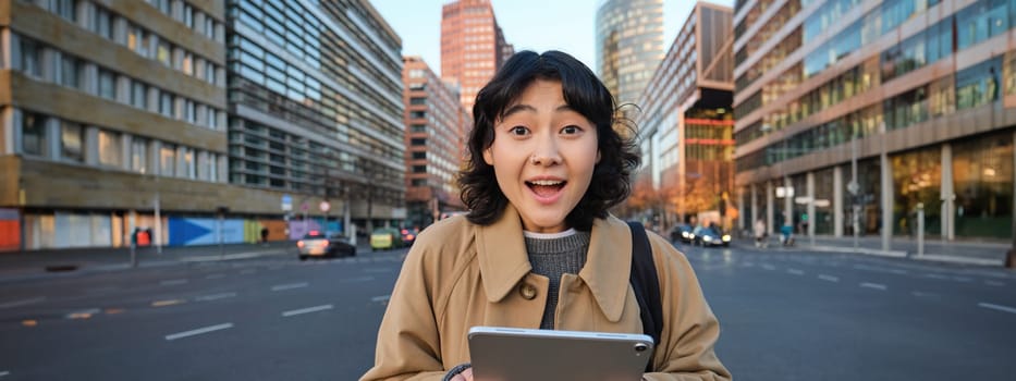 Portrait of korean girl looks surprised, found out something amazing, holding digital tablet with joyful face, stands on street of city centre.