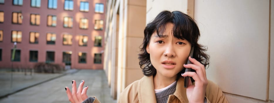 Korean girl with concerned face talks on mobile phone and shrugs, frowns and looks concerned, being lost in town, stands on street.