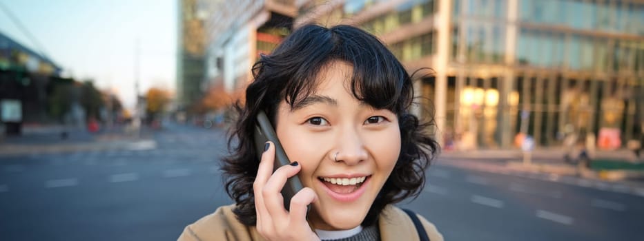 Portrait of happy korean girl, talks on mobile phone, looks surprised and happy, receives positive great news over telephone conversation, stands on street of city.
