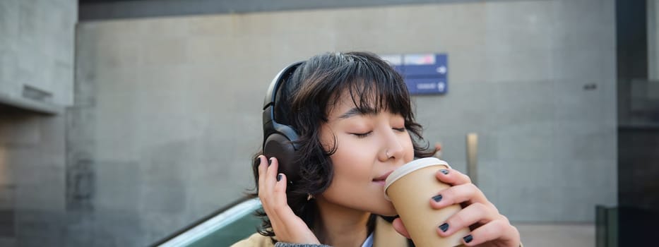 Happy brunette woman in headphones, listens music, enjoy drinking cup of coffee, stands on street, train station.