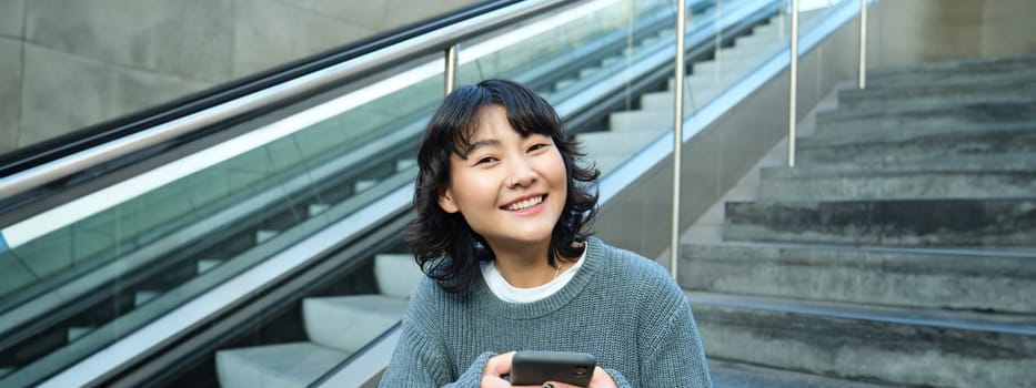 Portrait of brunette young woman, student sits on stairs in public place, looks at smartphone, reads text message and smiles.