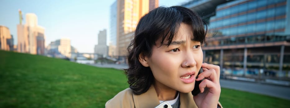 Close up portrait of young asian woman talks on mobile phone with concerned face, hear bad news over telephone conversation, walks from university with tablet.