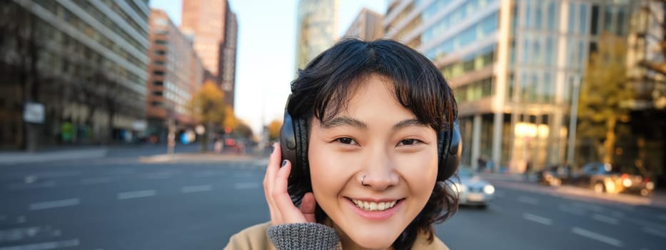 Portrait of young smiling korean girl, walking along city centre, listening music in headphones and holding mobile phone. Copy space