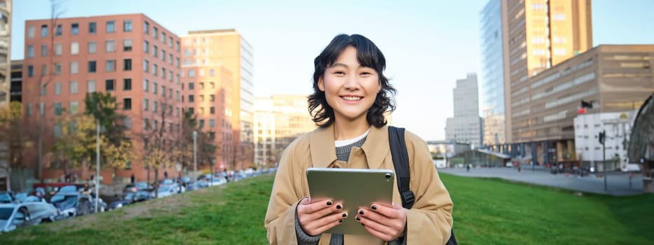 Happy asian girl stands on street, university student walks with digital tablet in hands and smiles, stands in city centre.