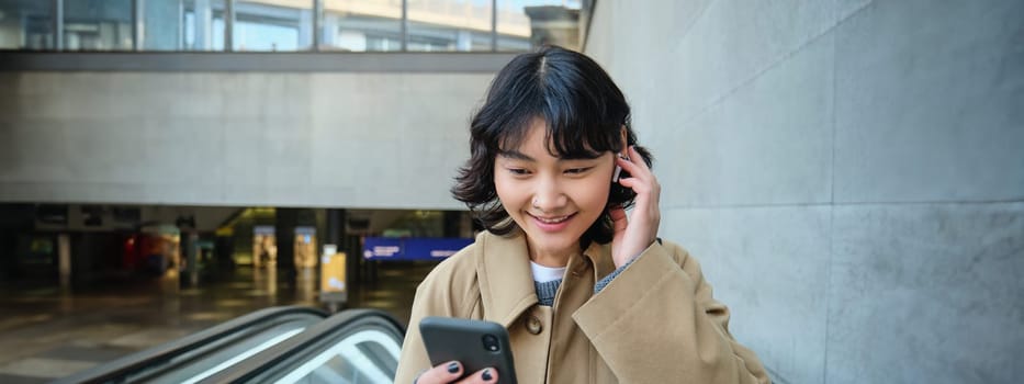 Portrait of cute korean girl in trench coat, going up escalator, holds mobile phone and smiles pleased, watches video in wireless headphones.