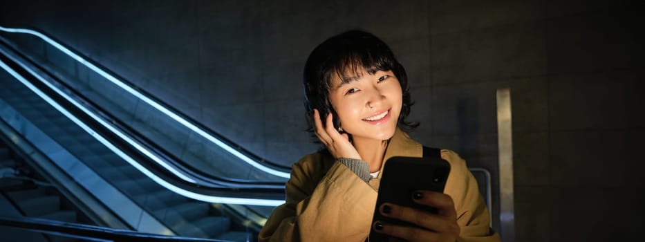 Portrait of beautiful korean girl in headphones, stands near escalator, travels, commutes home on public transport, holds smartphone, poses with mobile phone.