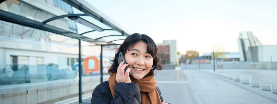 Beautiful smiling korean girl, waiting on bus stop, using public transport, talking on mobile phone, going somewhere in city.