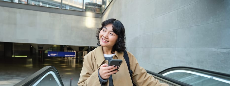 Smiling korean girl, student with smartphone goes up an escalator, reads mobile phone text message.