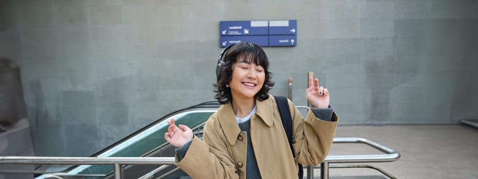 Happy young girl, student has fun on street, dances, listens music in headphones, drinks coffee. Copy space