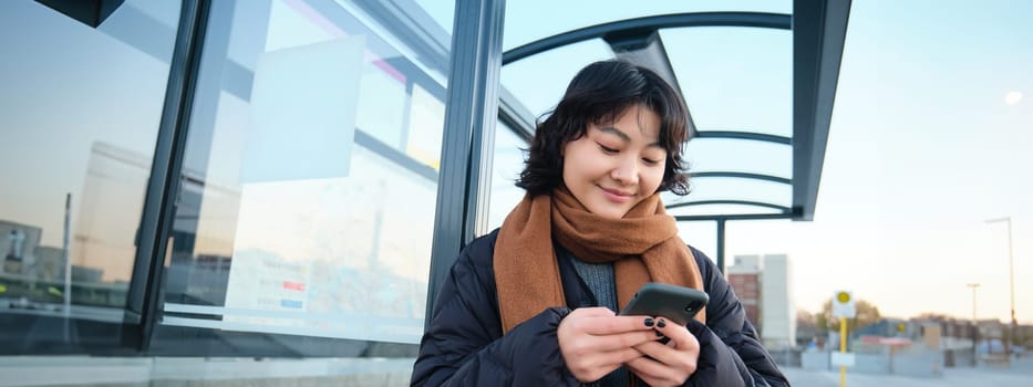 Beautiful Japanese girl, student standing on bus stop in winter jacket, holding smartphone, using application to track her public transport, ordering a ride.