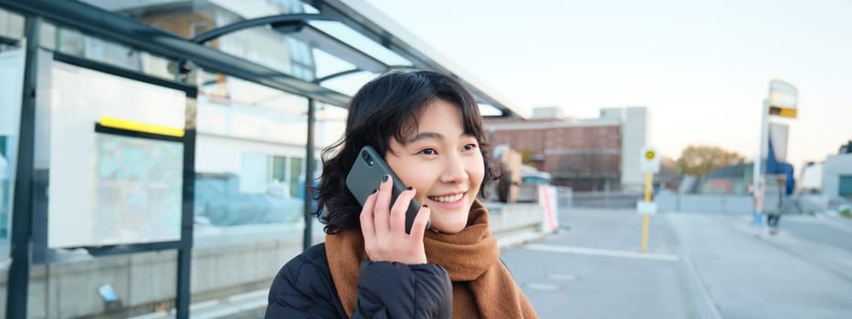 Smilling Korean girl talking on mobile phone, standing on bus stop, using smartphone, posing on road in winter, wrapped in scarf, wearing black jacket.