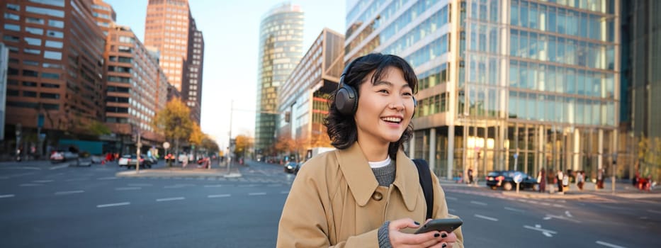 Portrait of young smiling korean girl, walking along city centre, listening music in headphones and holding mobile phone. Copy space