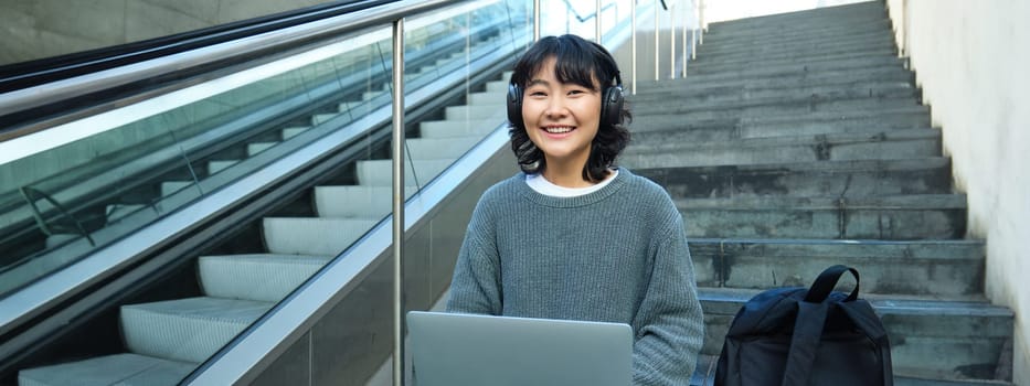 Female student works on project remotely, sits on stairs street, works on laptop, listens music in headphones and smiles.