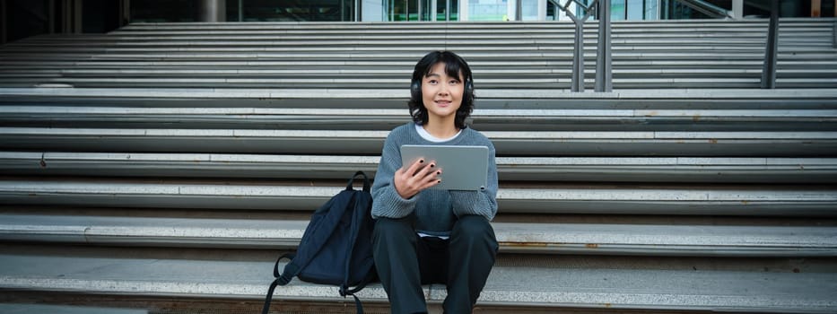 Young woman draws on her tablet, listens to music in headphones. Asian girl sits on stairs and does graphic design project, sits on street stairs.