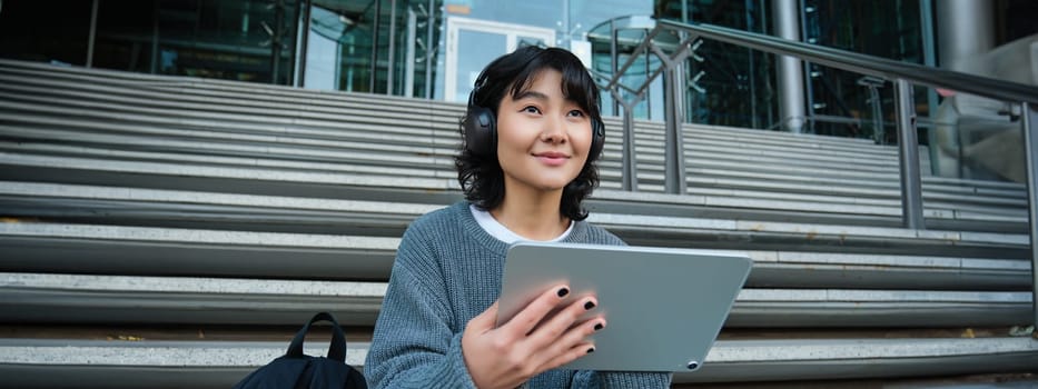 Young asian girl, student in headphones, works on remote, digital artist drawing on tablet with graphic pen, listening music in headphones and sitting on street staircase.