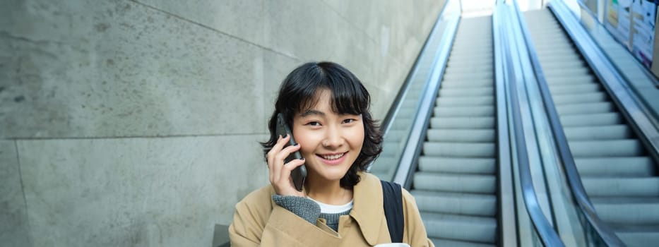 Portrait of smiling korean girl commutes, goes somewhere in city, drinks coffee to go and uses smartphone, stands on escalator.