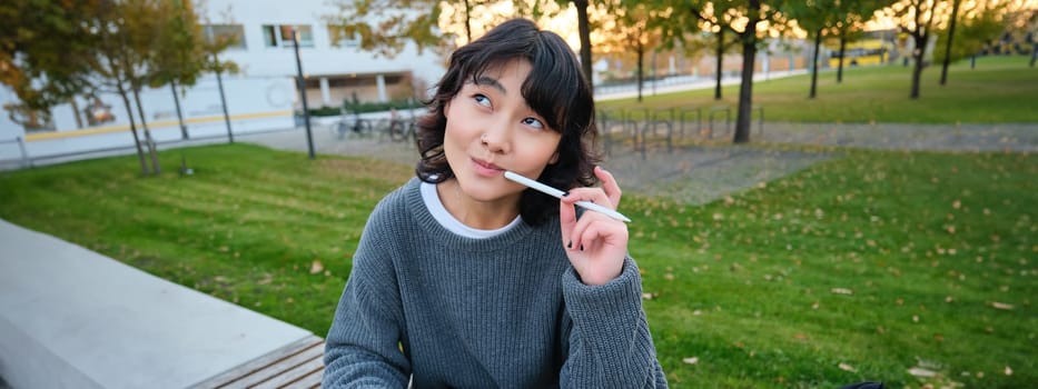 Portrait of asian woman, student in park, sits on bench with digital tablet and a pen, thinking, looking aside thoughtful, making notes, does her homework outdoors.