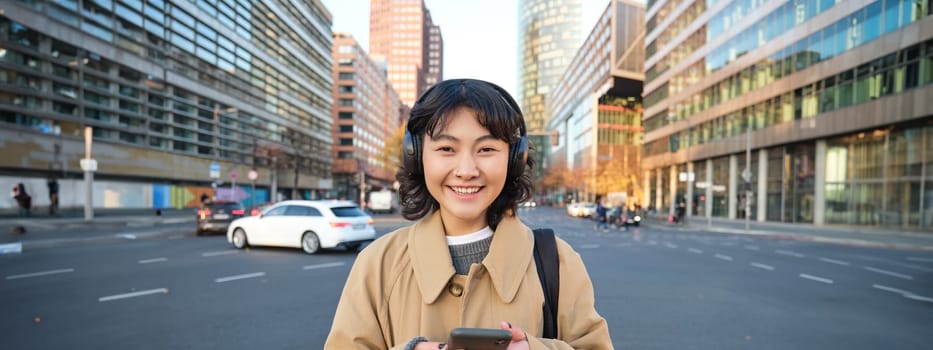 Portrait of young asian girl, student walks in city, listens music in headphones and uses mobile phone on streets.